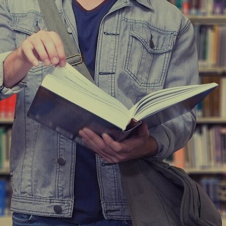 Eine Person mit einem Buch in der Hand steht vor einem Bibliotheksregal.